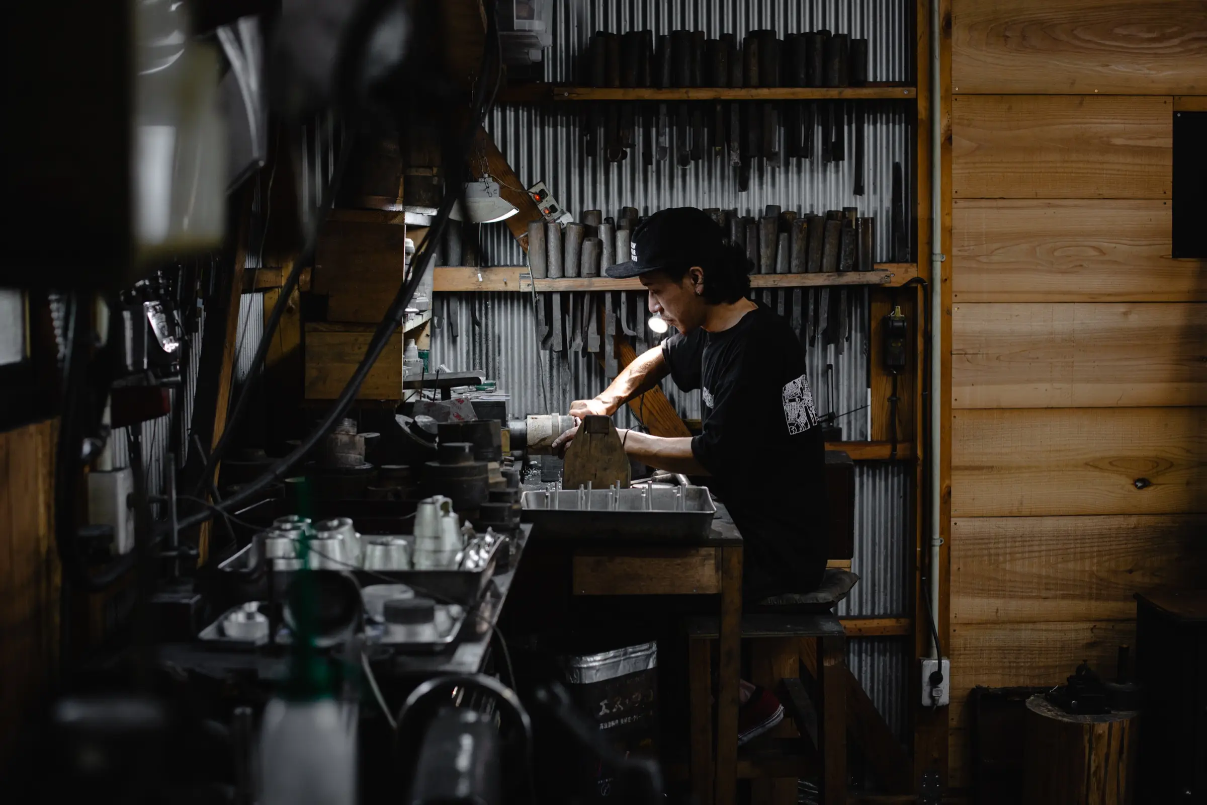 工具や錫の器が並ぶ薄暗い工房で、作業台に向かう錫師